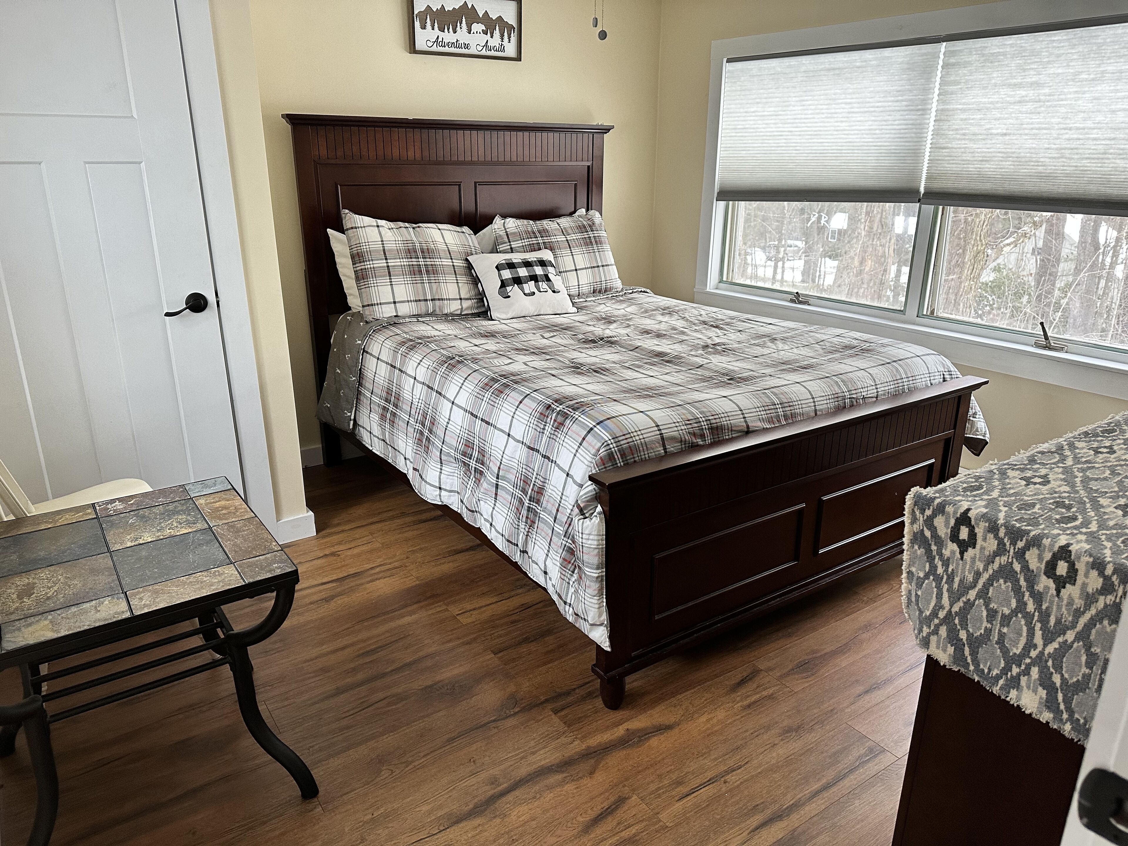 Une chambre à coucher avec un lit en bois, une couverture à carreaux, une fenêtre avec des stores et une petite table recouverte d’une nappe à motifs.