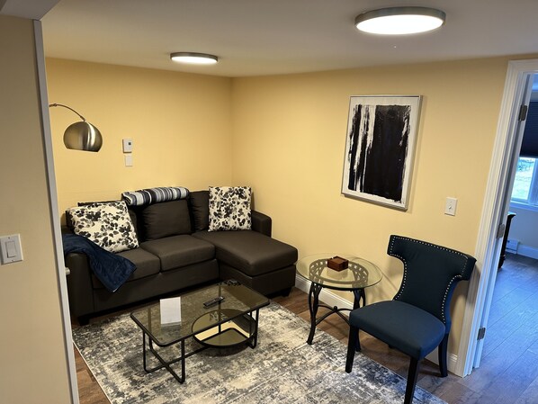 A living room with a sectional sofa, a glass coffee table, a small round table, and a dark armchair.