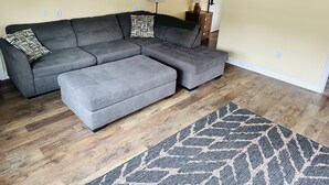 Un salon avec un canapé d’angle gris, un miroir rond, une lampe et un tapis à motifs.