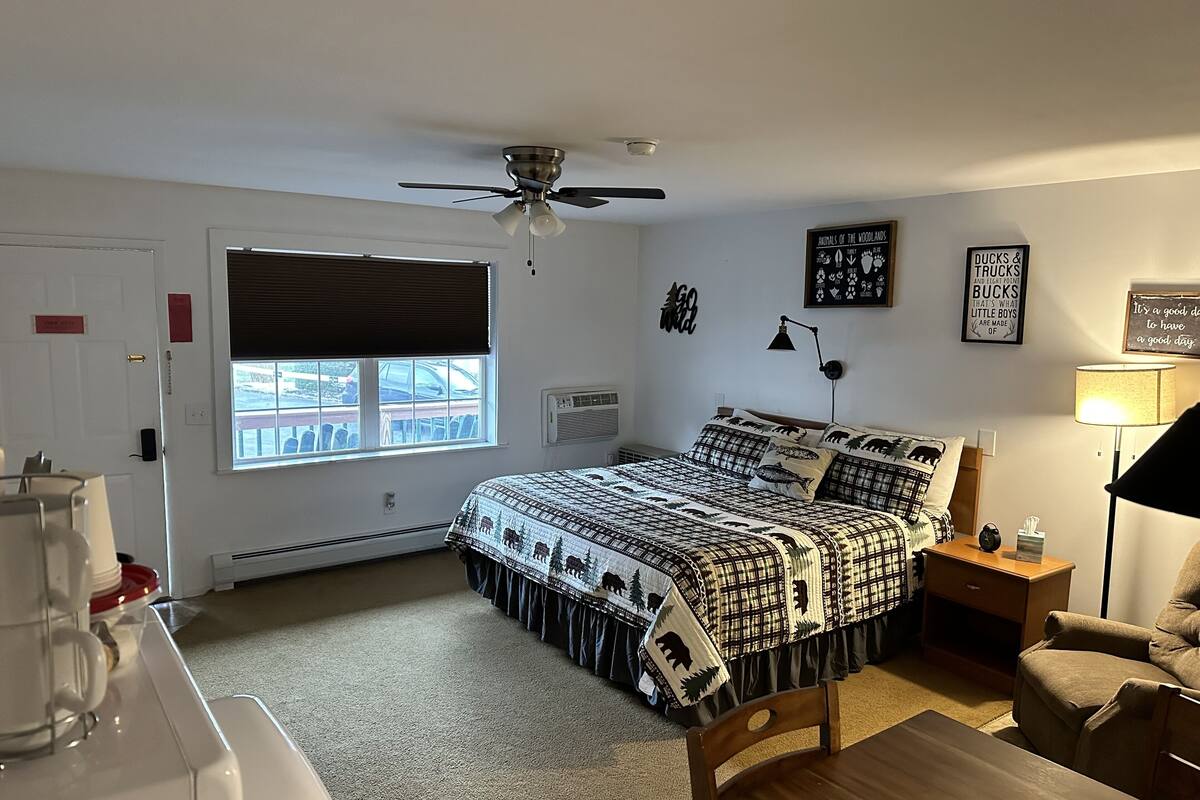 A bedroom with a bed, a desk, a chair, a ceiling fan, a window, and a wall with framed pictures.