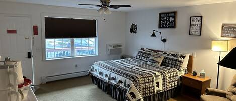 A bedroom with a bed, a desk, a chair, a ceiling fan, a window, and a wall with framed pictures.
