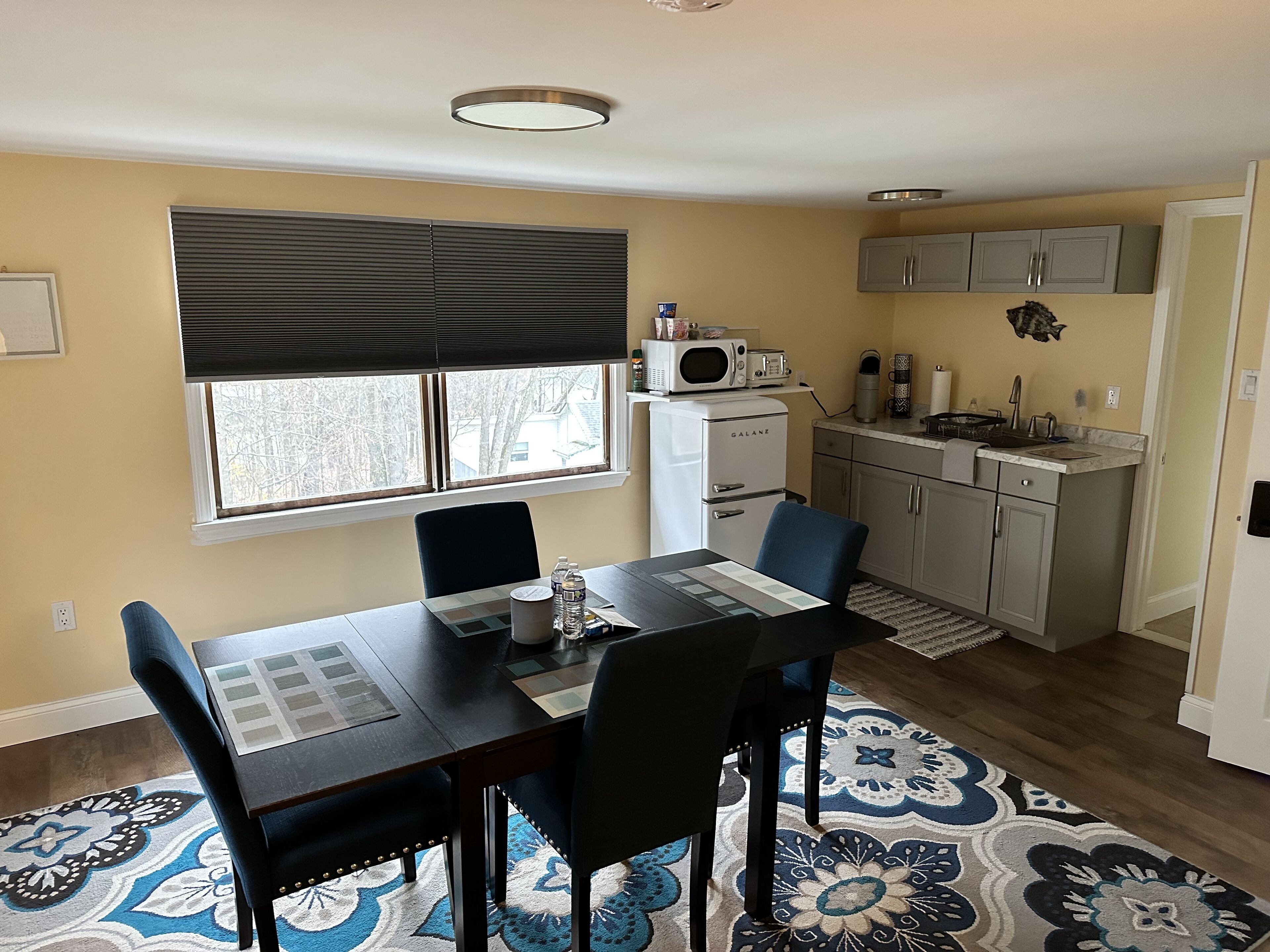 A compact kitchen with a refrigerator, microwave, and coffee maker. There is a dining table with chairs and a patterned rug.