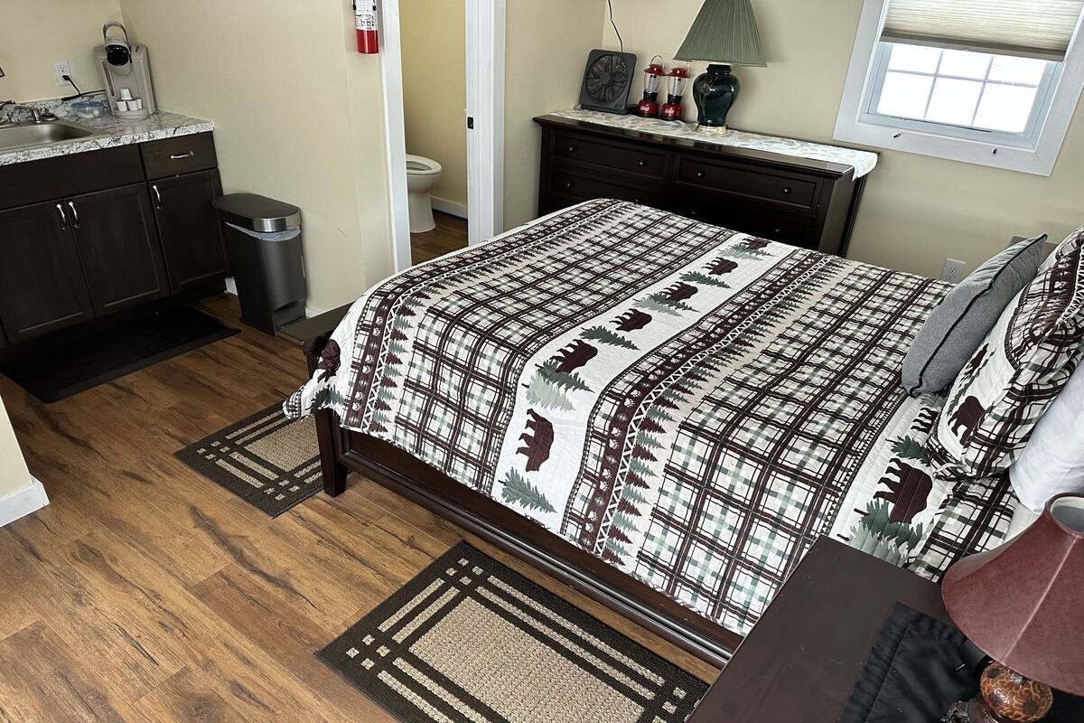 A bedroom with a bed, a dresser, a nightstand, a window, and a bathroom visible through an open door.