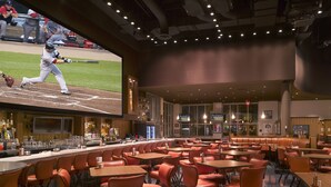 A sports bar with a large screen displaying a baseball game, multiple seating areas with tables and chairs, and a bar with a variety of drinks. - Northern Quest Resort And Casino (Airway Heights)