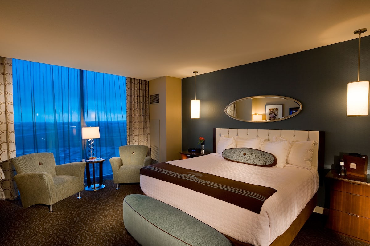 A hotel room with a large bed, two armchairs, a side table, a lamp, a mirror, and a view of the ocean.