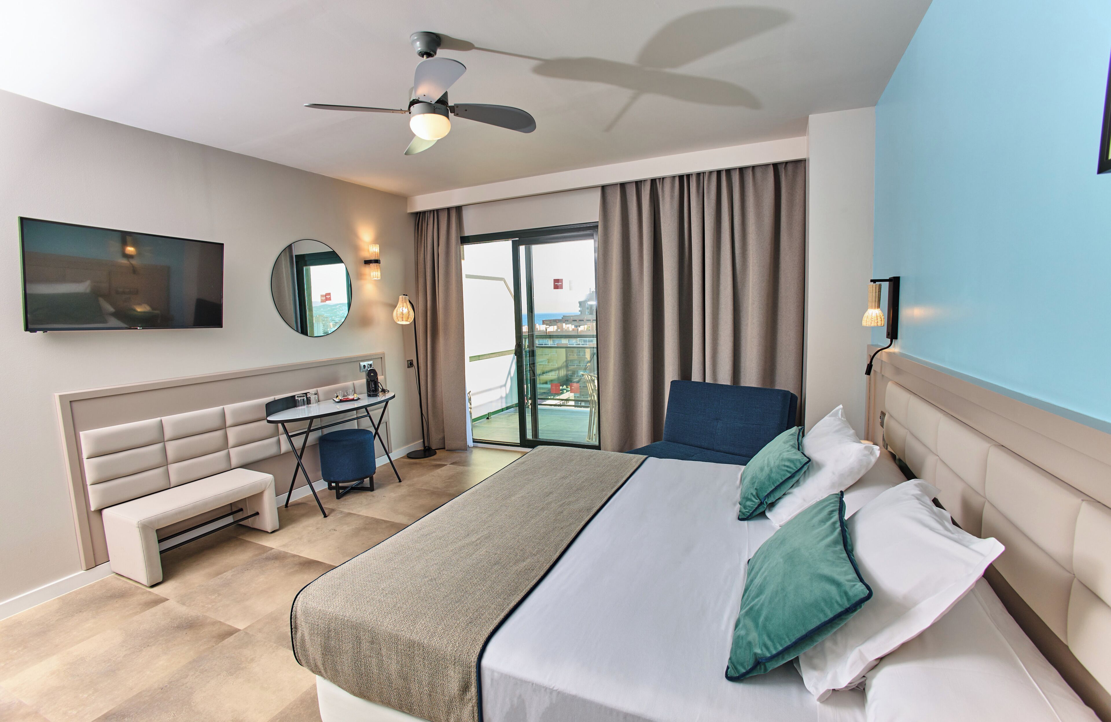 Une chambre d’hôtel moderne dotée d’un grand lit, d’un bureau, d’une chaise, d’une banquette, d’une télévision, d’un ventilateur de plafond et offrant une vue sur l’extérieur.