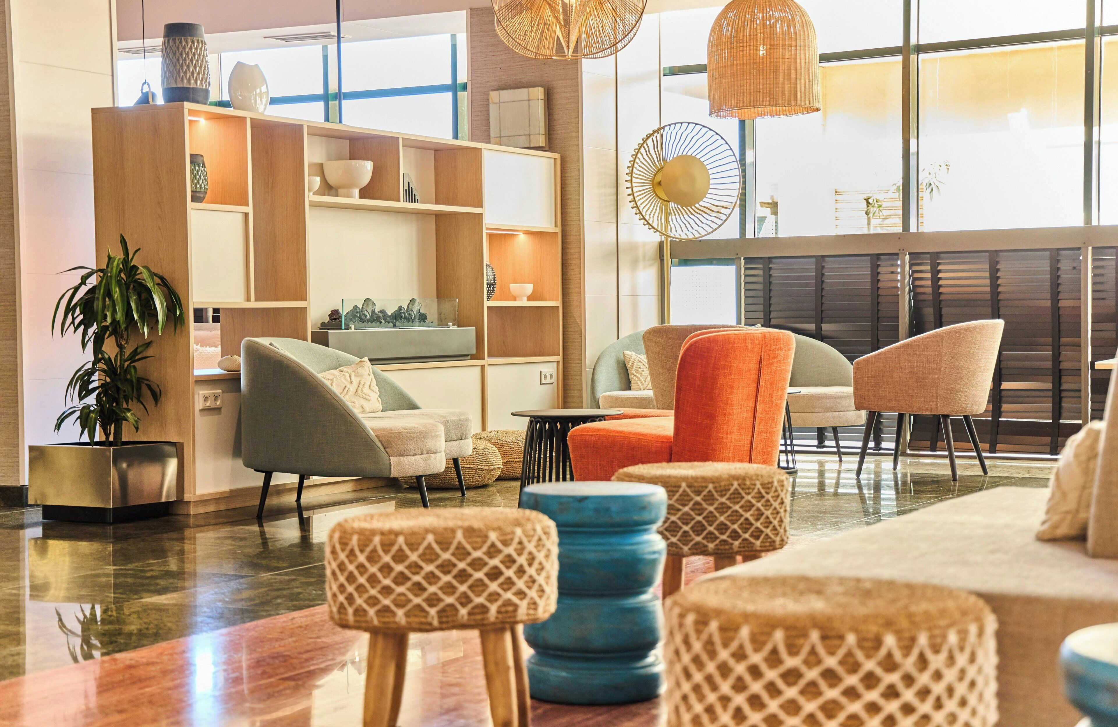 A modern lounge area with a wooden bookshelf, comfortable seating, a potted plant, and a large window.