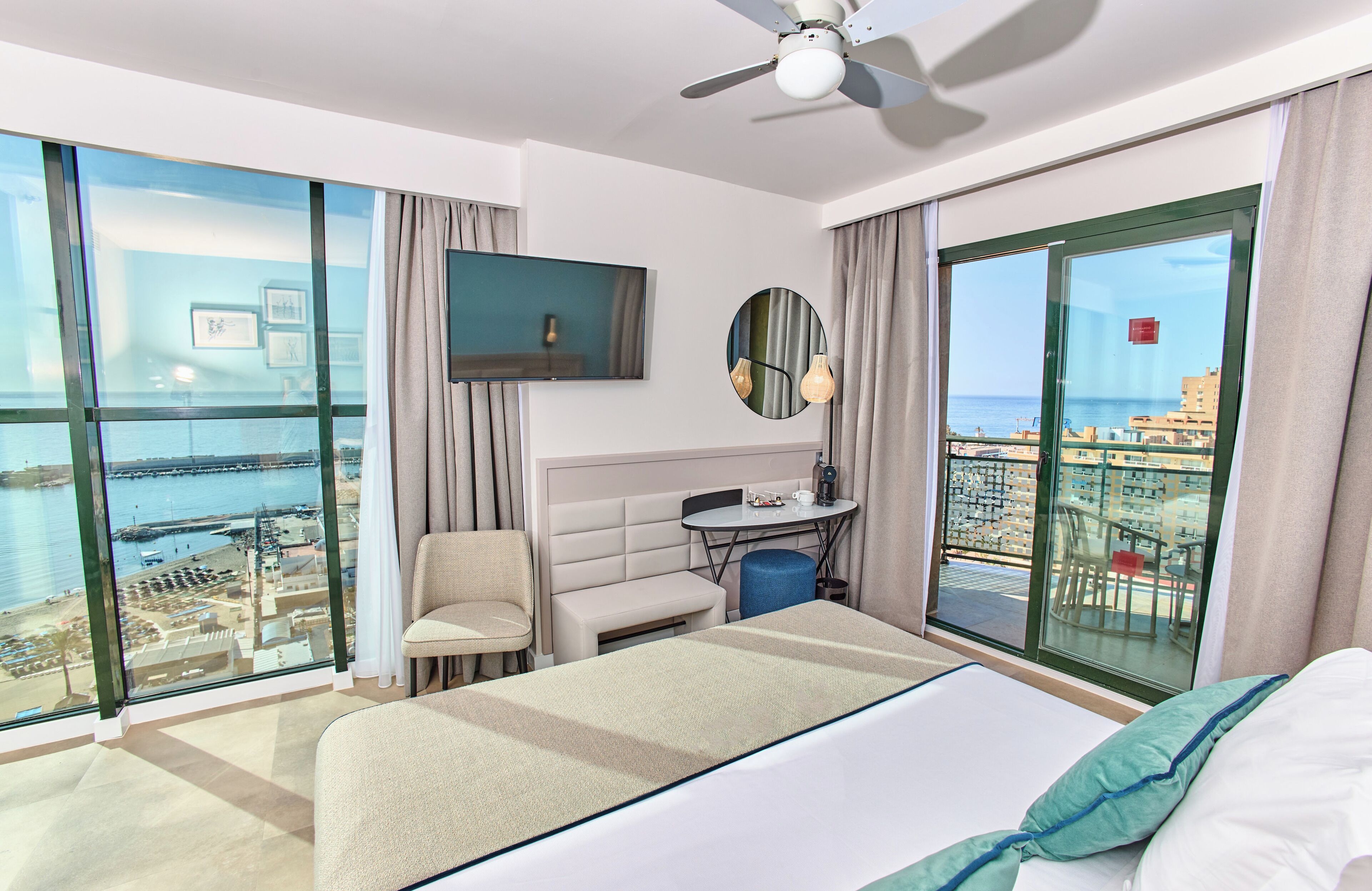 A hotel room with a large bed, a seating area, a small table, a TV, and a balcony with a view of the sea and buildings.