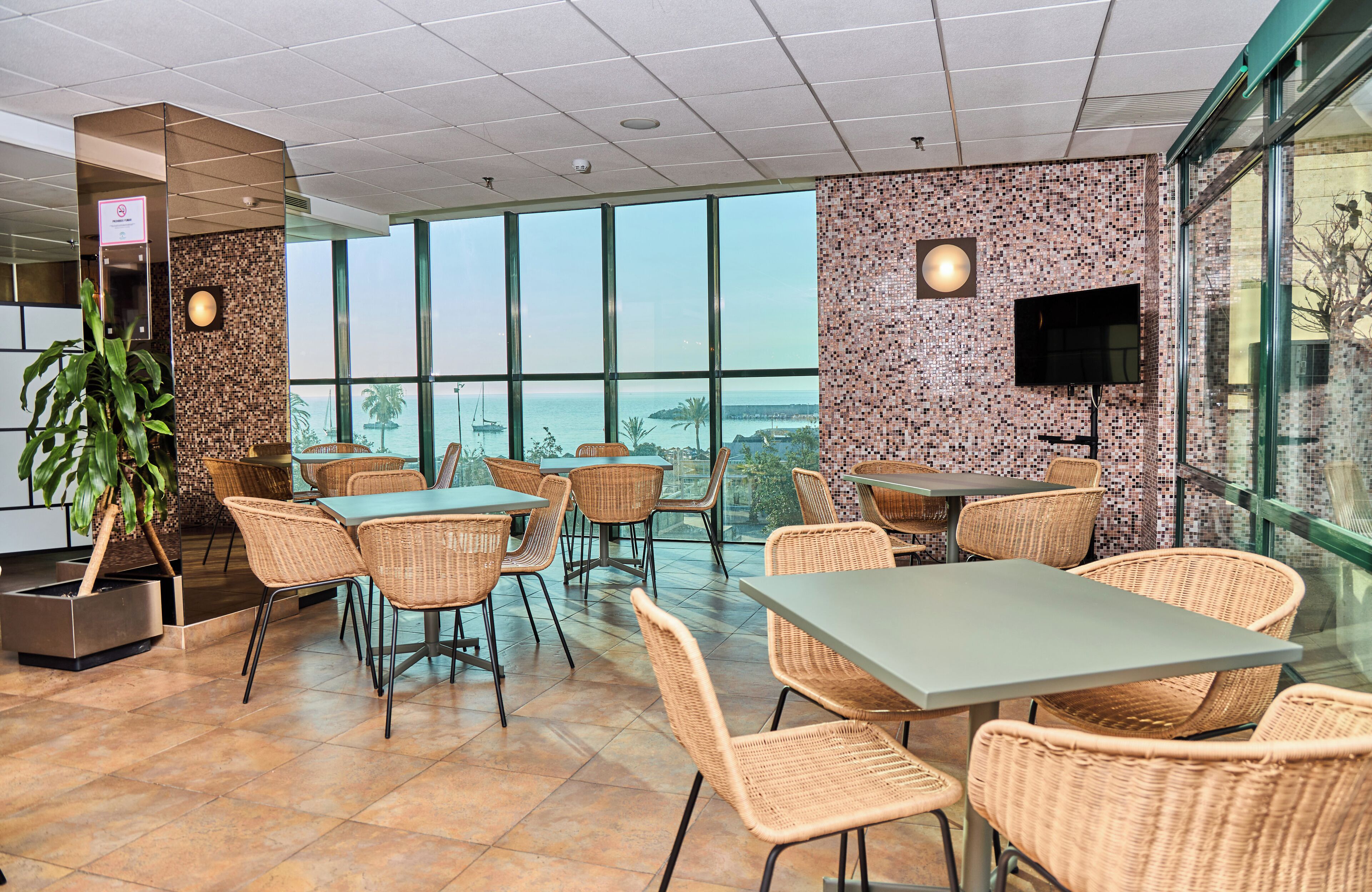 A restaurant with wicker chairs and tables, large windows with a view of the ocean, and a flat-screen TV mounted on the wall.