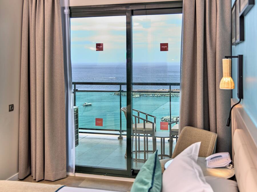 Une chambre d’hôtel dotée d’une grande porte-fenêtre coulissante donnant sur un balcon avec vue sur la mer et un quai. Le balcon est équipé de deux chaises et d’une table, et la chambre comprend un lit avec un oreiller et une télécommande.