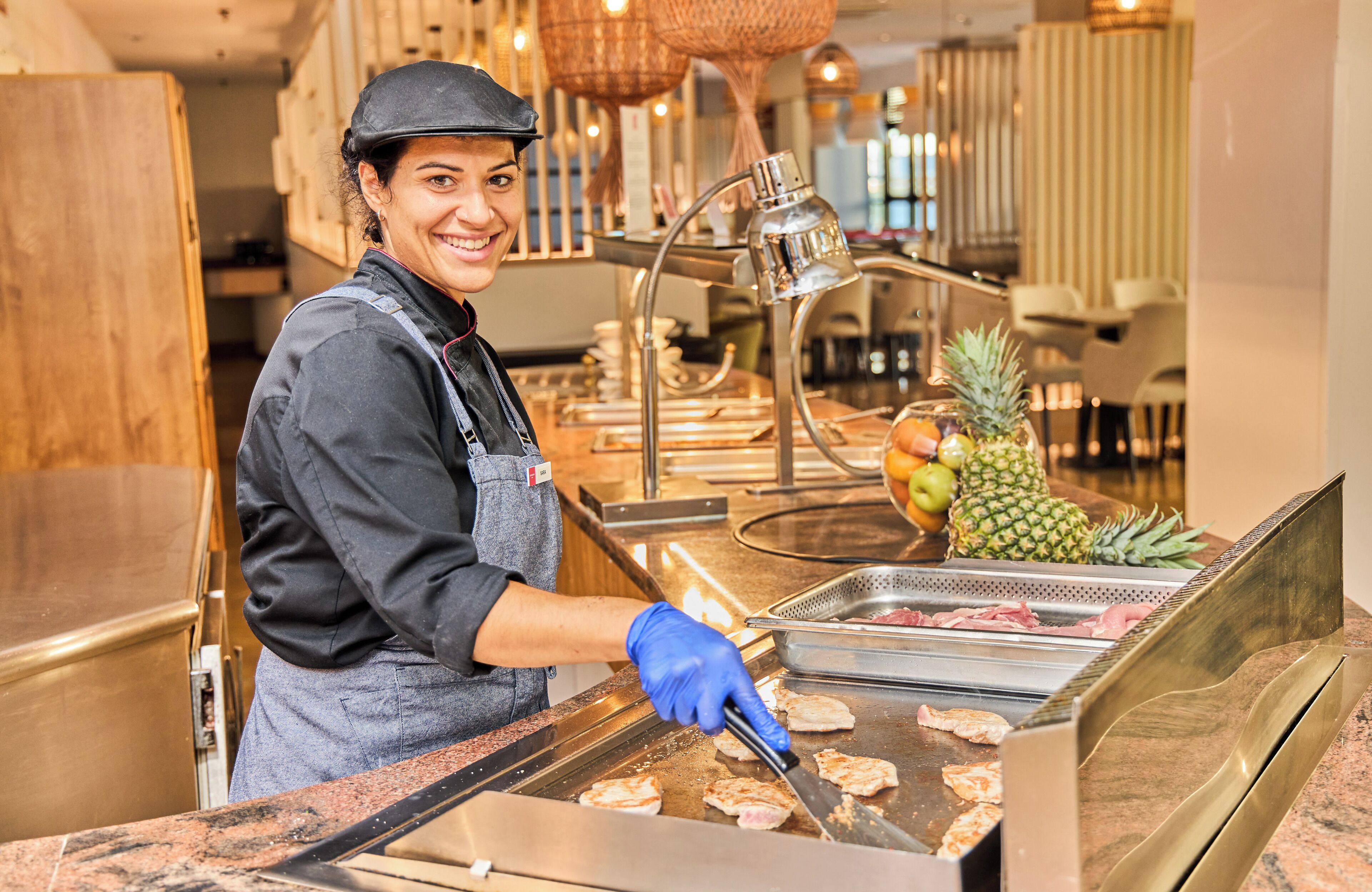 Una persona cucina al banco di un buffet con una griglia, un ananas e una ciotola di frutta sullo sfondo.