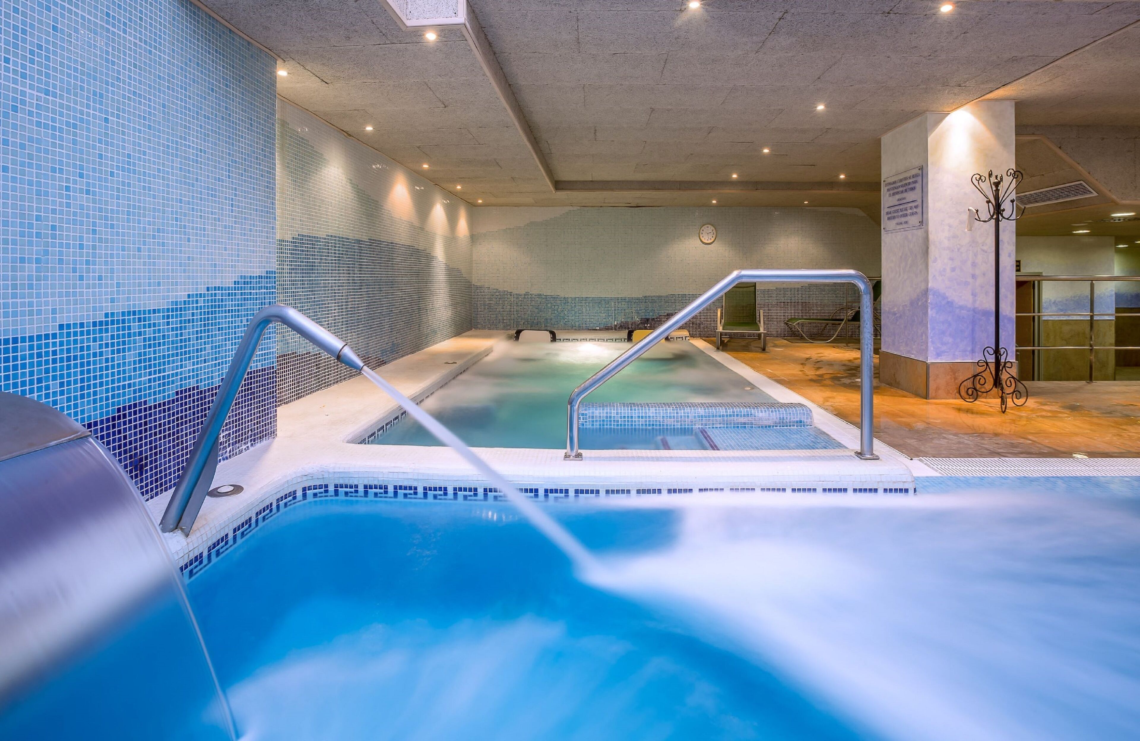 An indoor swimming pool with a tiled wall, a hot tub, and a lounge area with a chair.