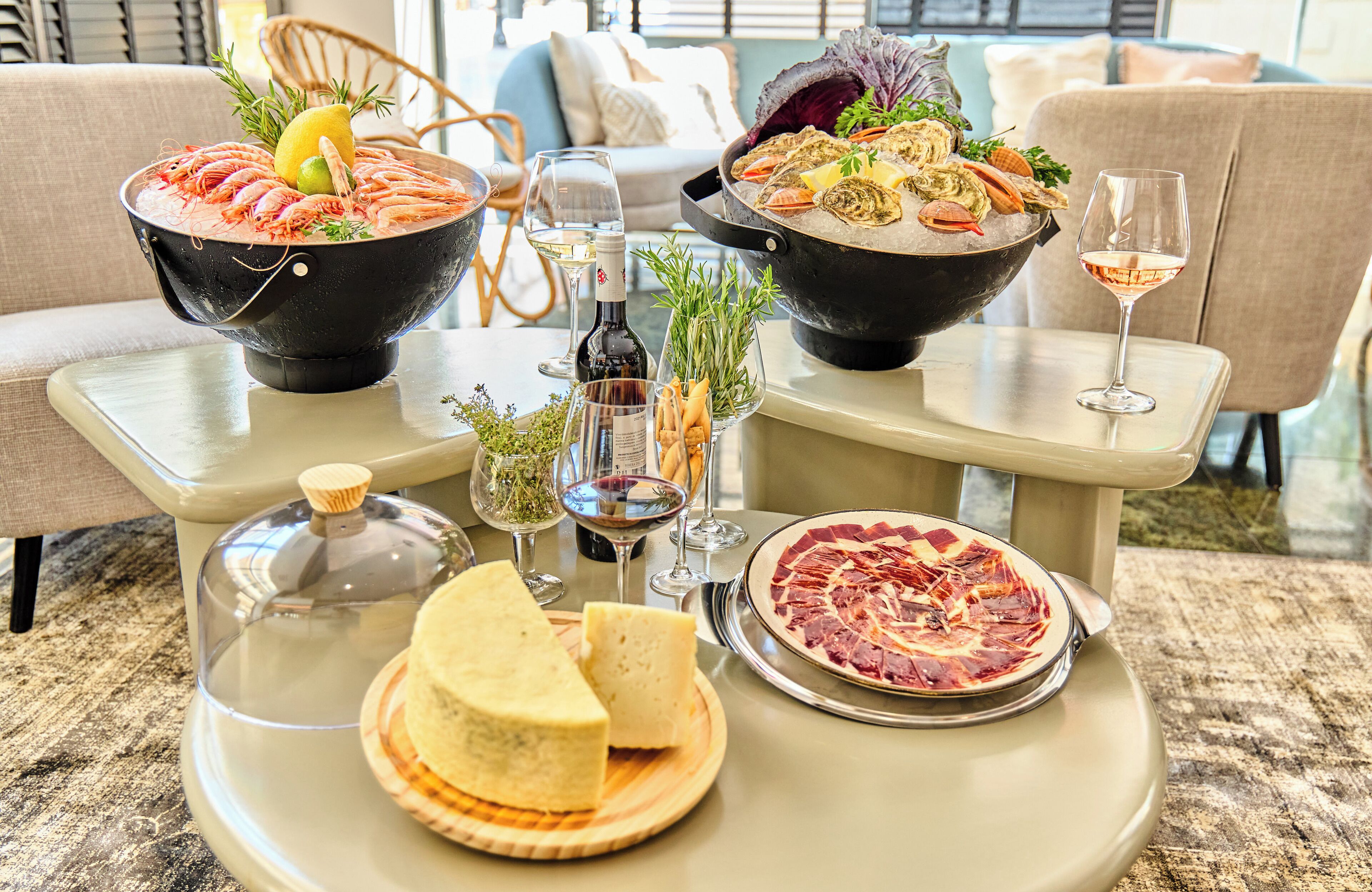 Une table à manger dressée avec des plats de fruits de mer, des verres à vin et des assiettes de fromage, dans une pièce bien éclairée où l’on aperçoit un canapé et des fauteuils au fond.