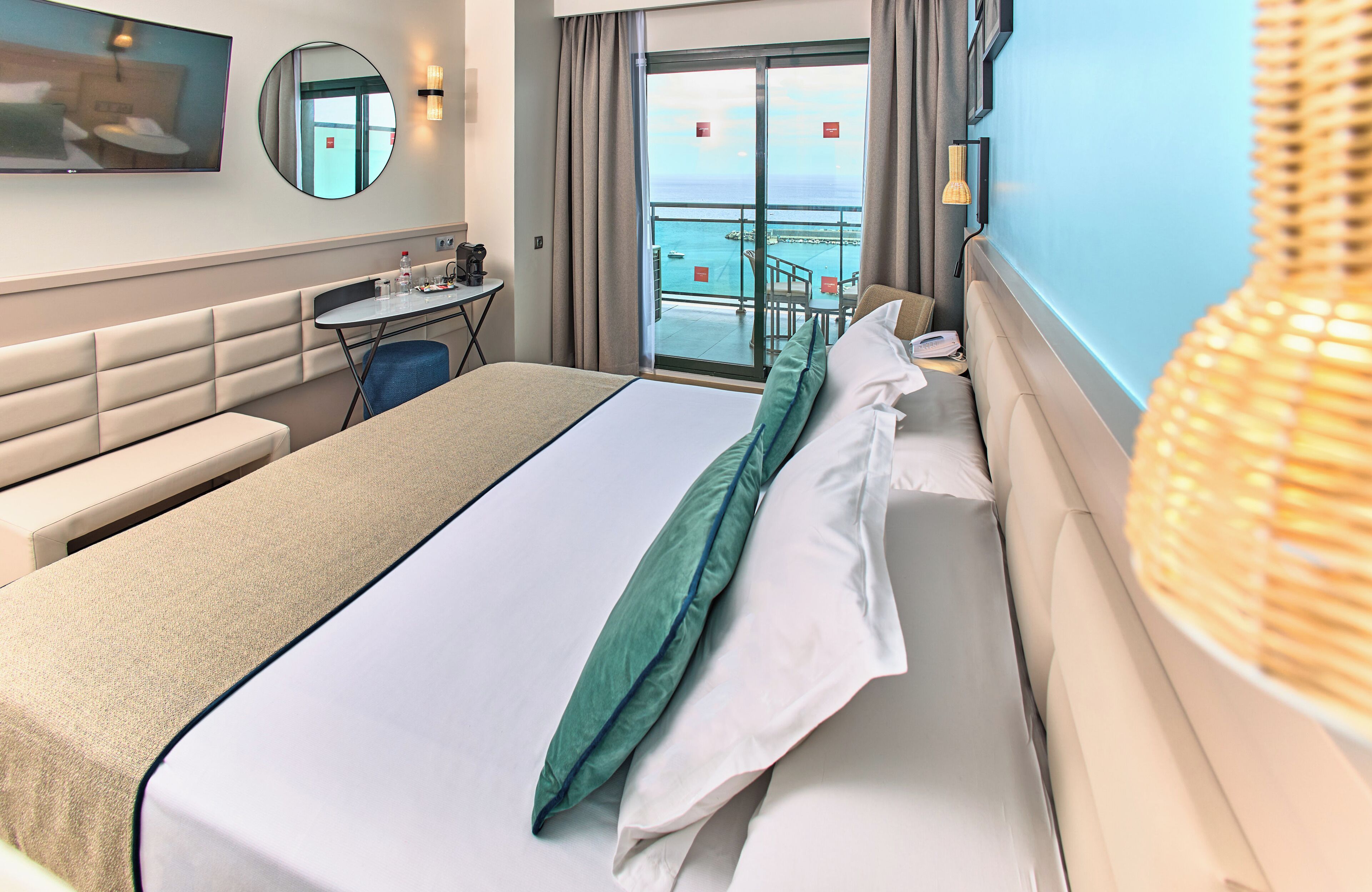 Ein Hotelzimmer mit einem großen Bett, einem Schreibtisch, einem Sessel, einem kleinen Tisch, einem Spiegel und Meerblick durch eine Schiebetür.