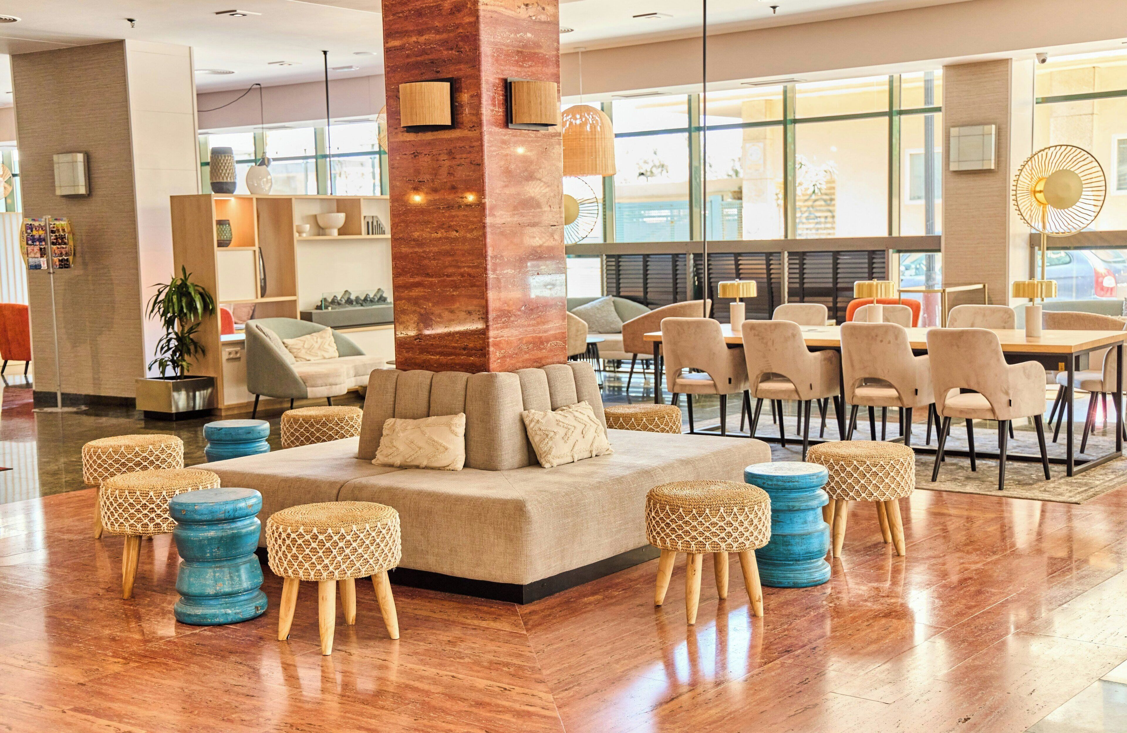 A modern hotel lobby with a central seating area, a large pillar, and a dining area with chairs and tables.