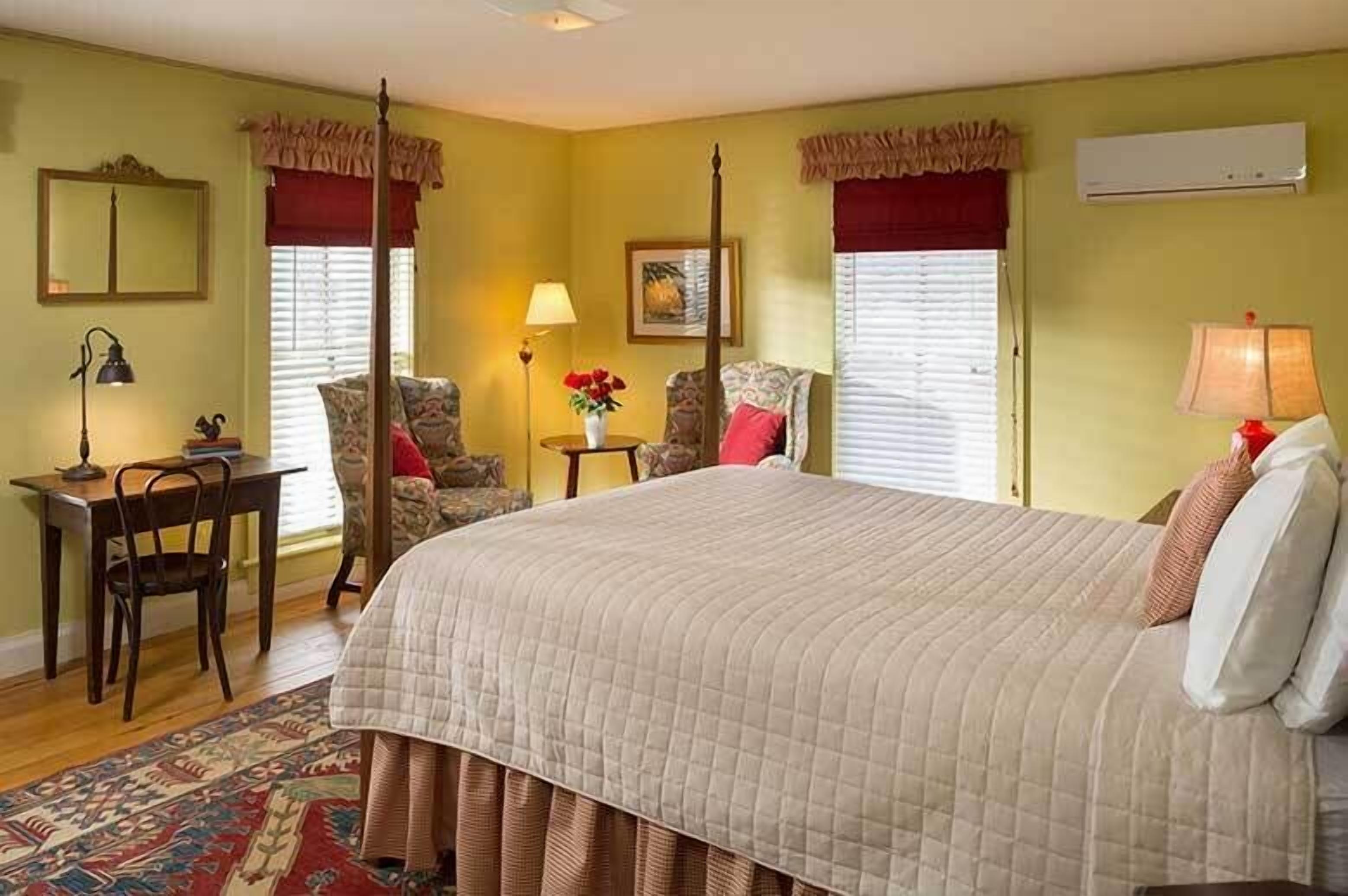 A bedroom with a large bed, a desk, and two chairs. There is a window with a curtain, a wall-mounted air conditioner, and a painting on the wall.