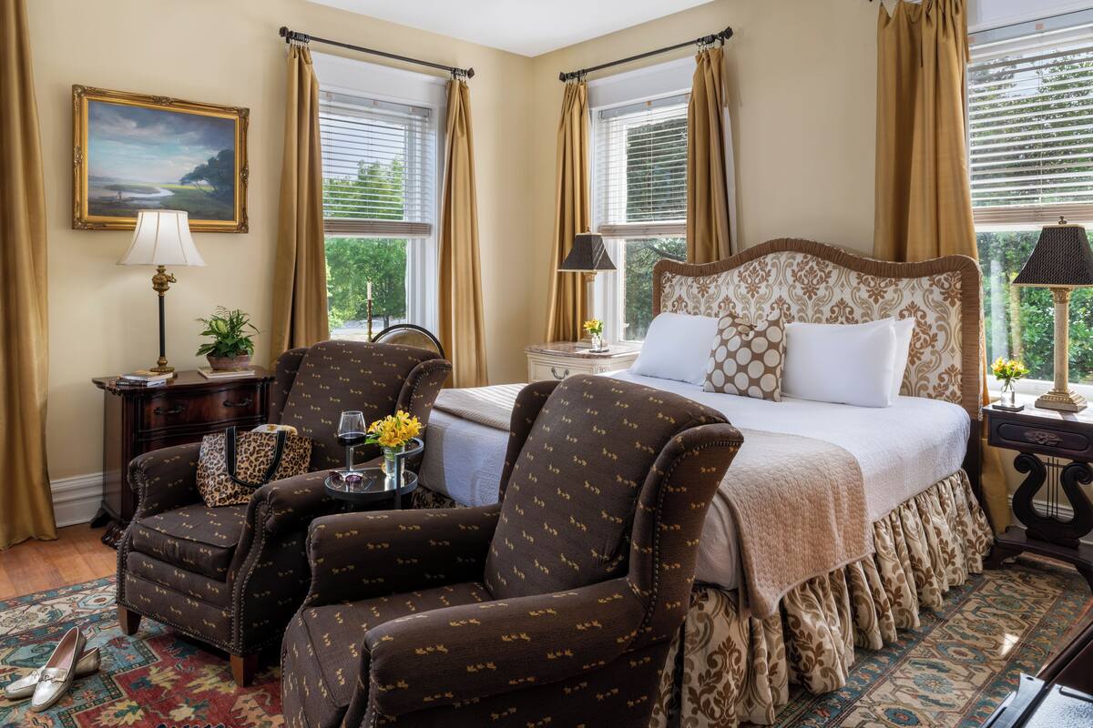 Quarto com cama grande, duas poltronas, mesa de cabeceira com luminárias, um quadro na parede e janela com cortinas.