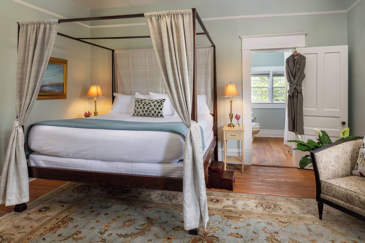 Uma cama com dossel e cortina, com edredom branco e azul, duas luminárias de cabeceira, uma mesinha de cabeceira, uma cadeira e vista para o exterior através de uma porta.