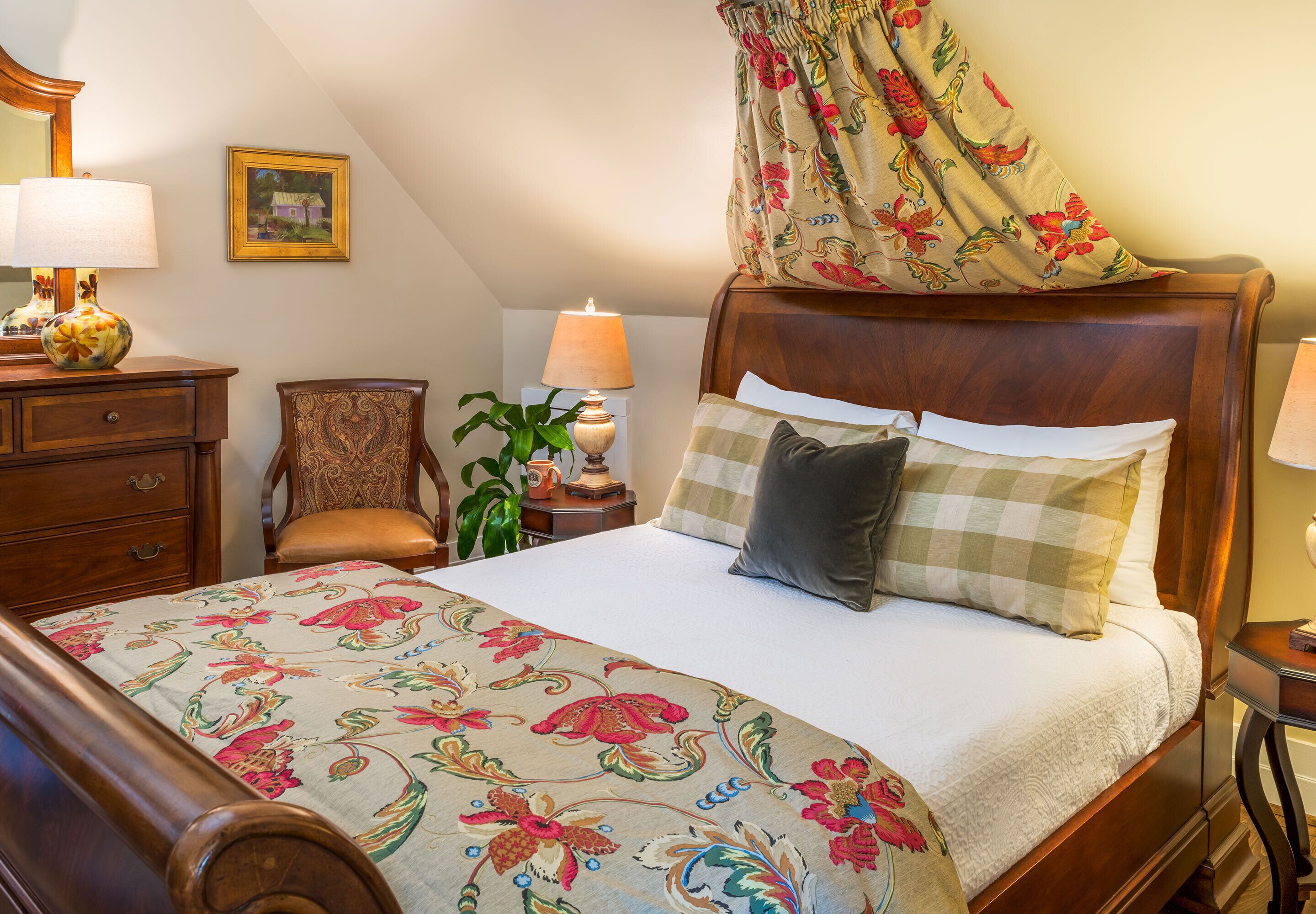 A bedroom with a wooden bed, floral bedspread, a chair, a nightstand with lamps, and a painting on the wall.
