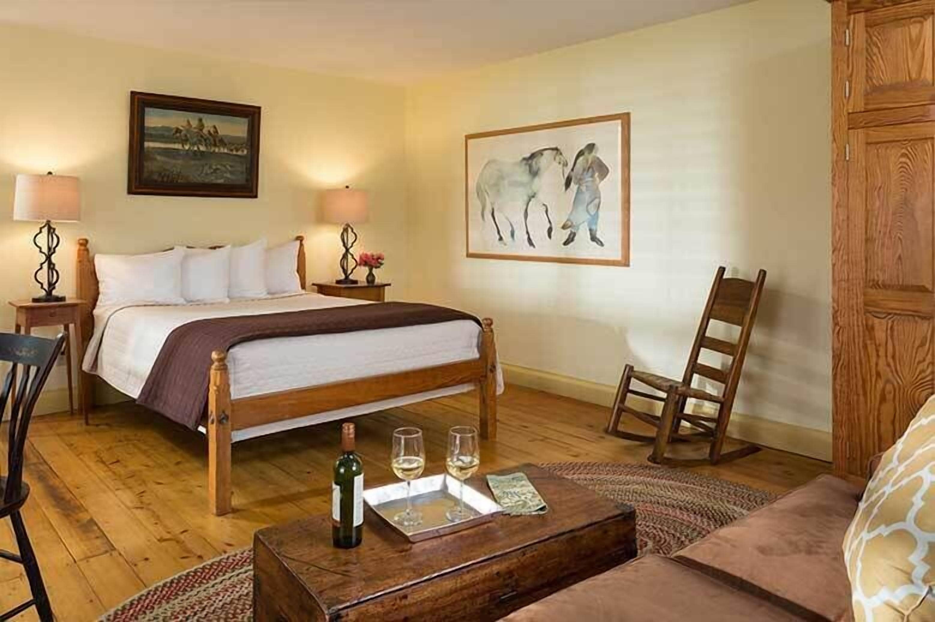 A bedroom with a wooden bed, two bedside lamps, a framed painting, a wooden chair, and a small table with a bottle and glasses.