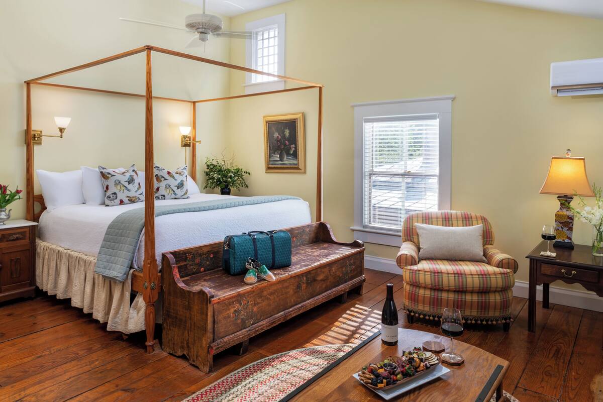 Uma cama com dossel de madeira, um banco, uma poltrona xadrez e uma mesa pequena com uma garrafa e copos.
