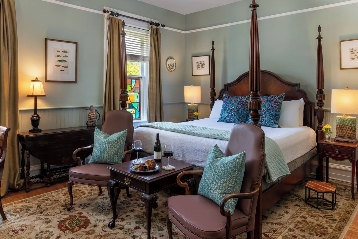 Quarto com cama de dossel, duas cadeiras, uma mesa com uma travessa de fruta e uma mesinha de cabeceira com uma garrafa de vinho e copos.