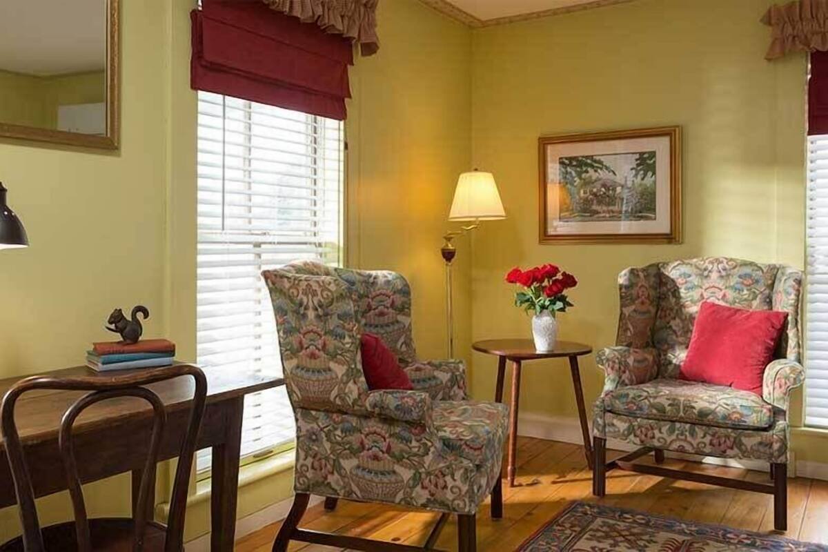 Quarto com duas poltronas estampadas, uma mesinha com um vaso de flores, um abajur e um quadro pendurado na parede.
