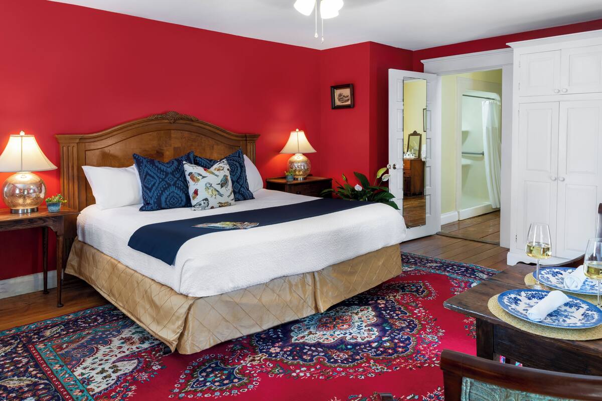 Quarto com uma cama grande, duas luminárias de cabeceira, parede vermelha, cabeceira de madeira, tapete estampado e guarda-roupa branco.