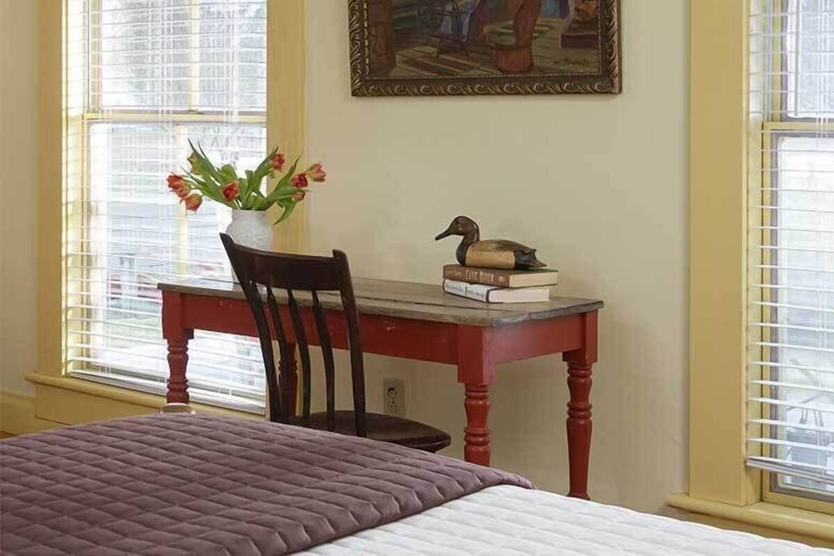 Quarto com cama, escrivaninha com cadeira, vaso com flores e quadro na parede.