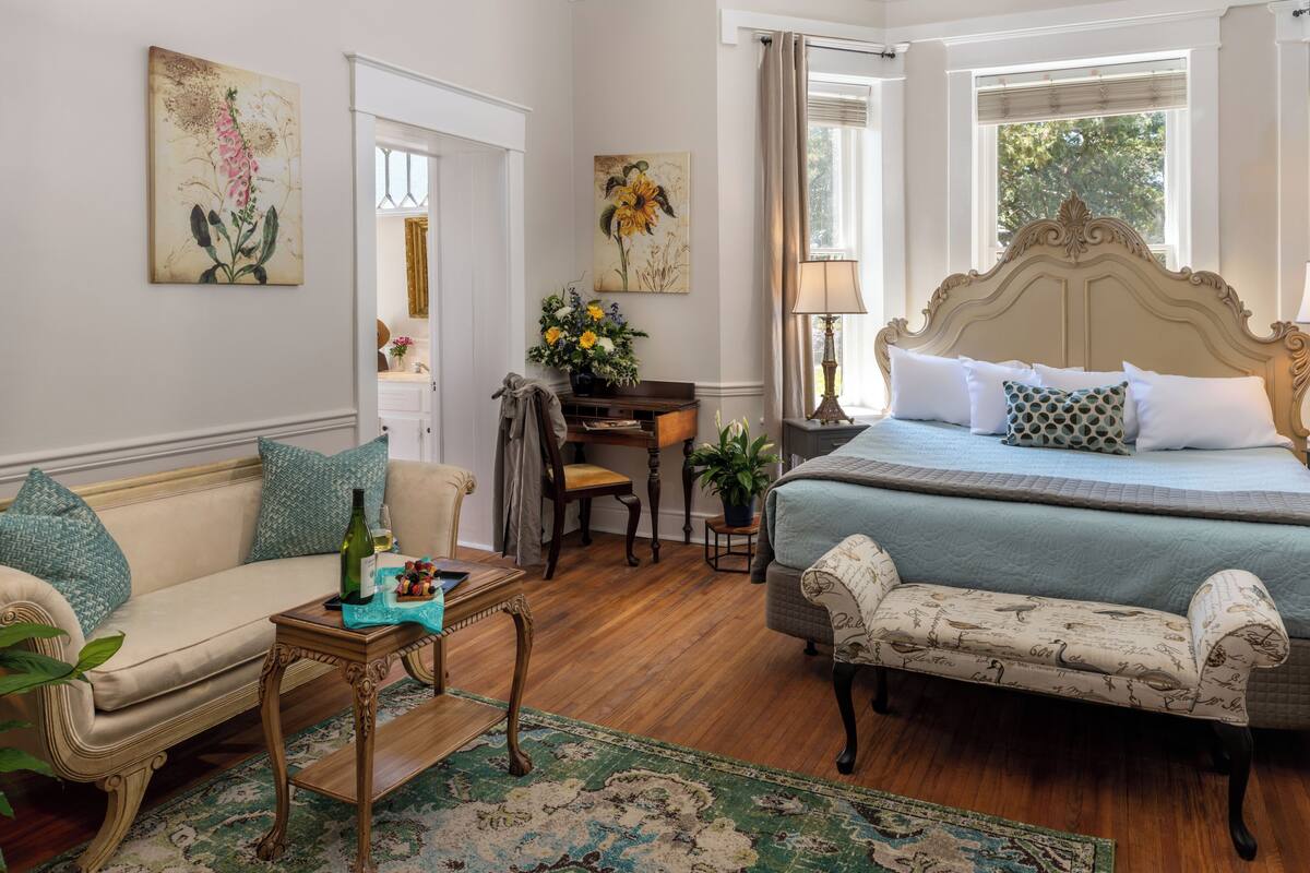 Quarto com cama, sofá, cadeira, mesa pequena com uma garrafa e duas pinturas na parede.