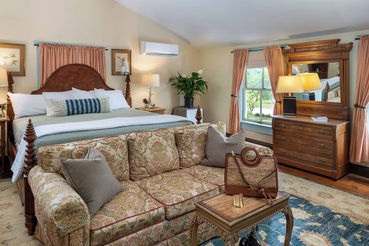 Quarto de hotel com cama grande, sofá, cômoda de madeira e janela com cortinas.