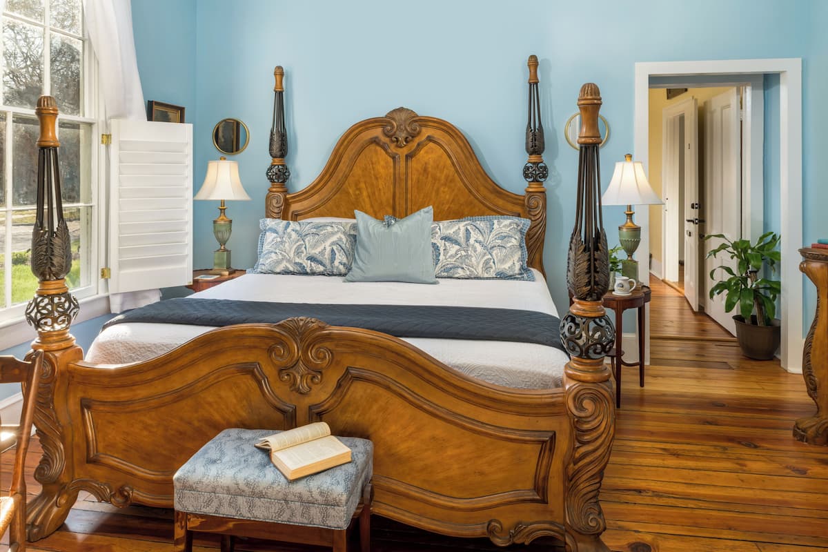 Quarto com uma cama de madeira grande, duas mesas de cabeceira com luminárias, uma janela com cortinas e vista para o corredor.