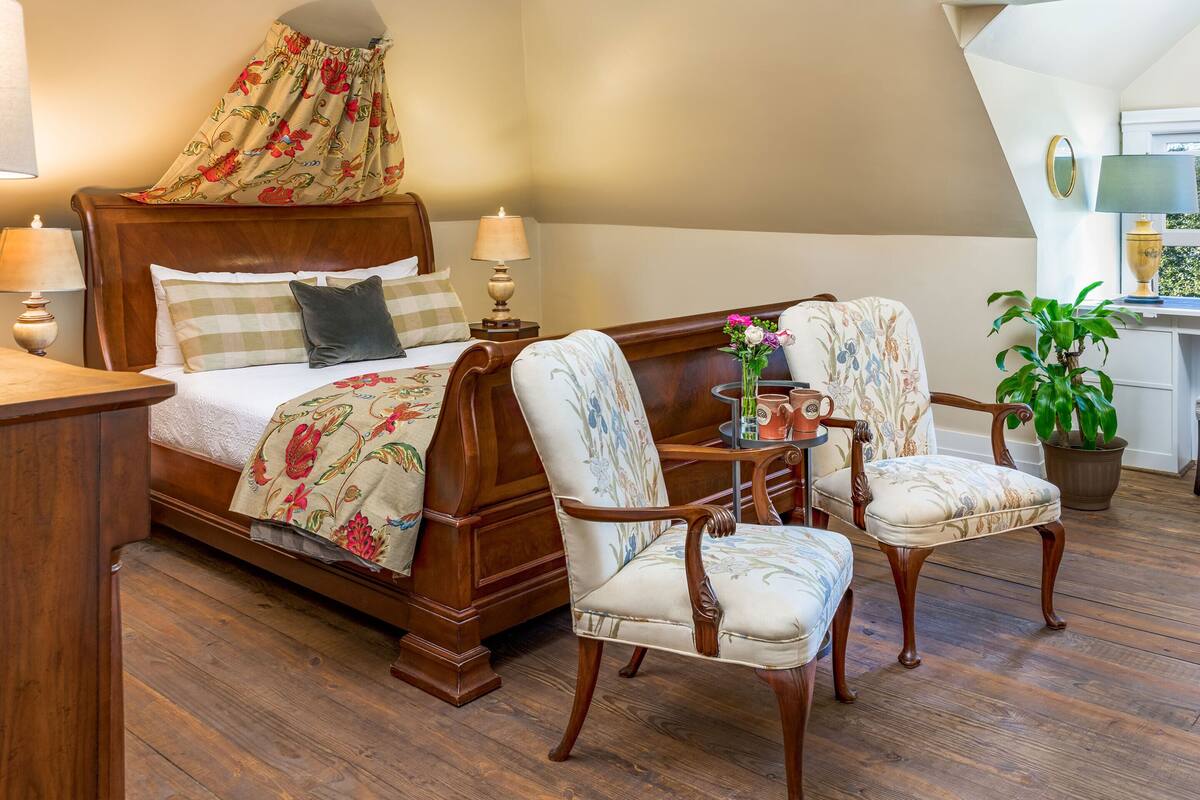 Quarto com cama de madeira, duas poltronas estofadas, uma mesinha de centro com um vaso de flores e um vaso de planta.