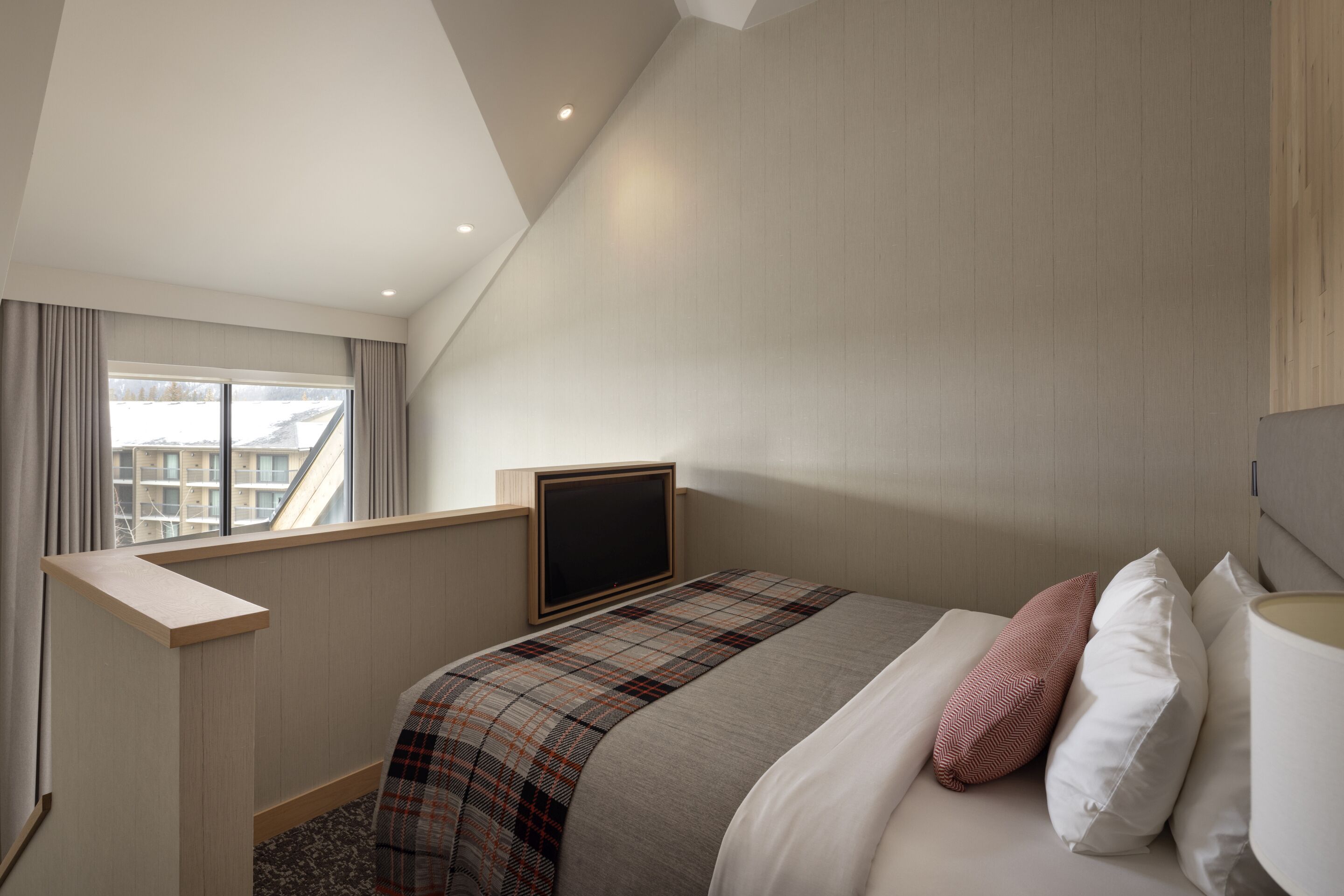 Un dormitorio con una cama, un televisor montado en la pared, una ventana con cortinas y vistas a edificios.