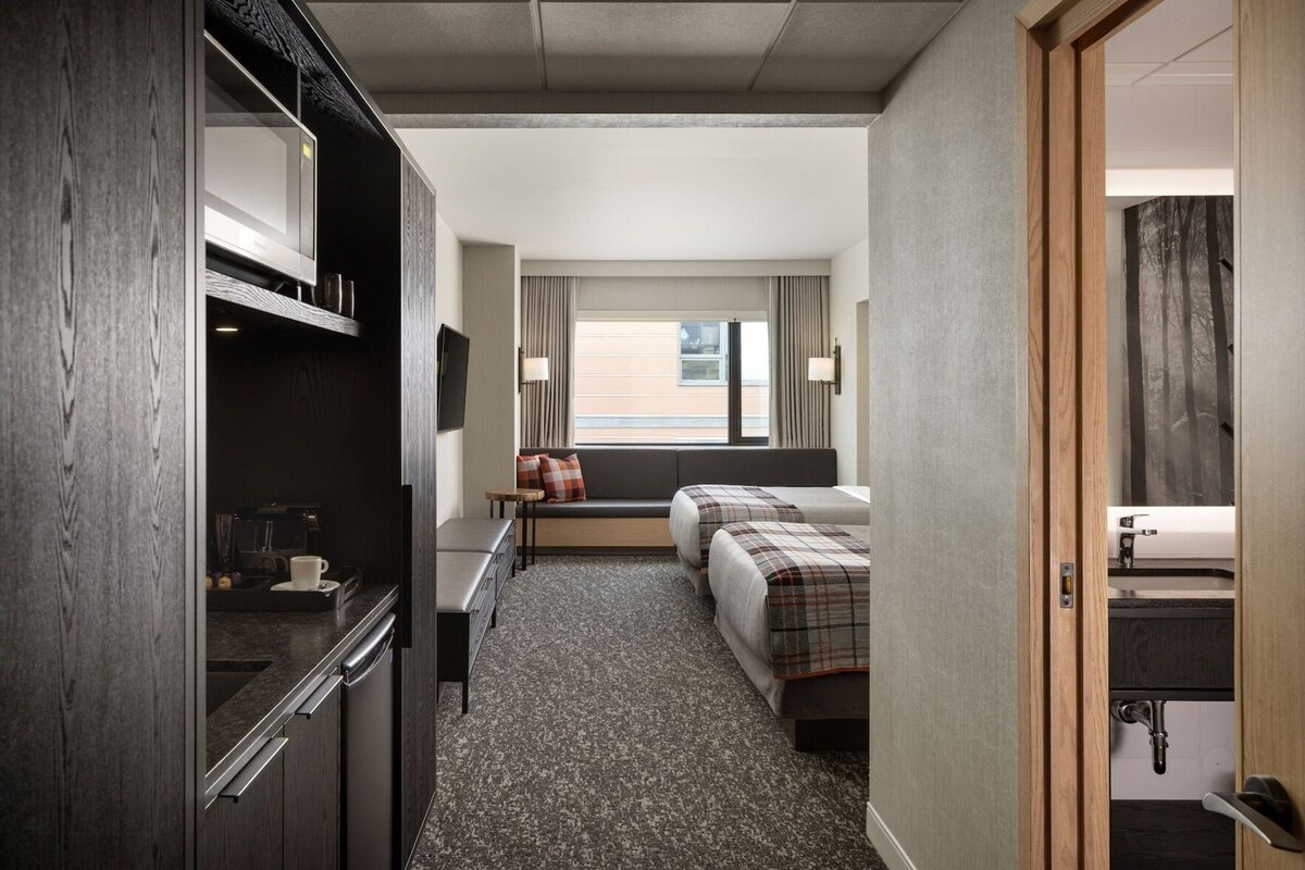 A hotel room with a kitchenette, a sofa, two beds, a window with curtains, and a view of the outside.
