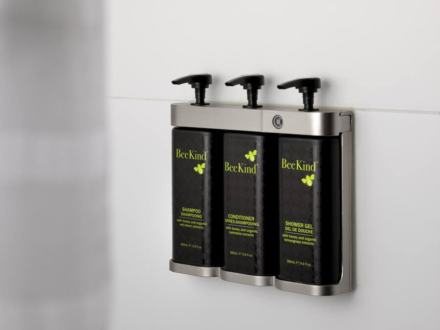 Trois bouteilles noires de produits BeeKind sont disposées sur un distributeur mural.