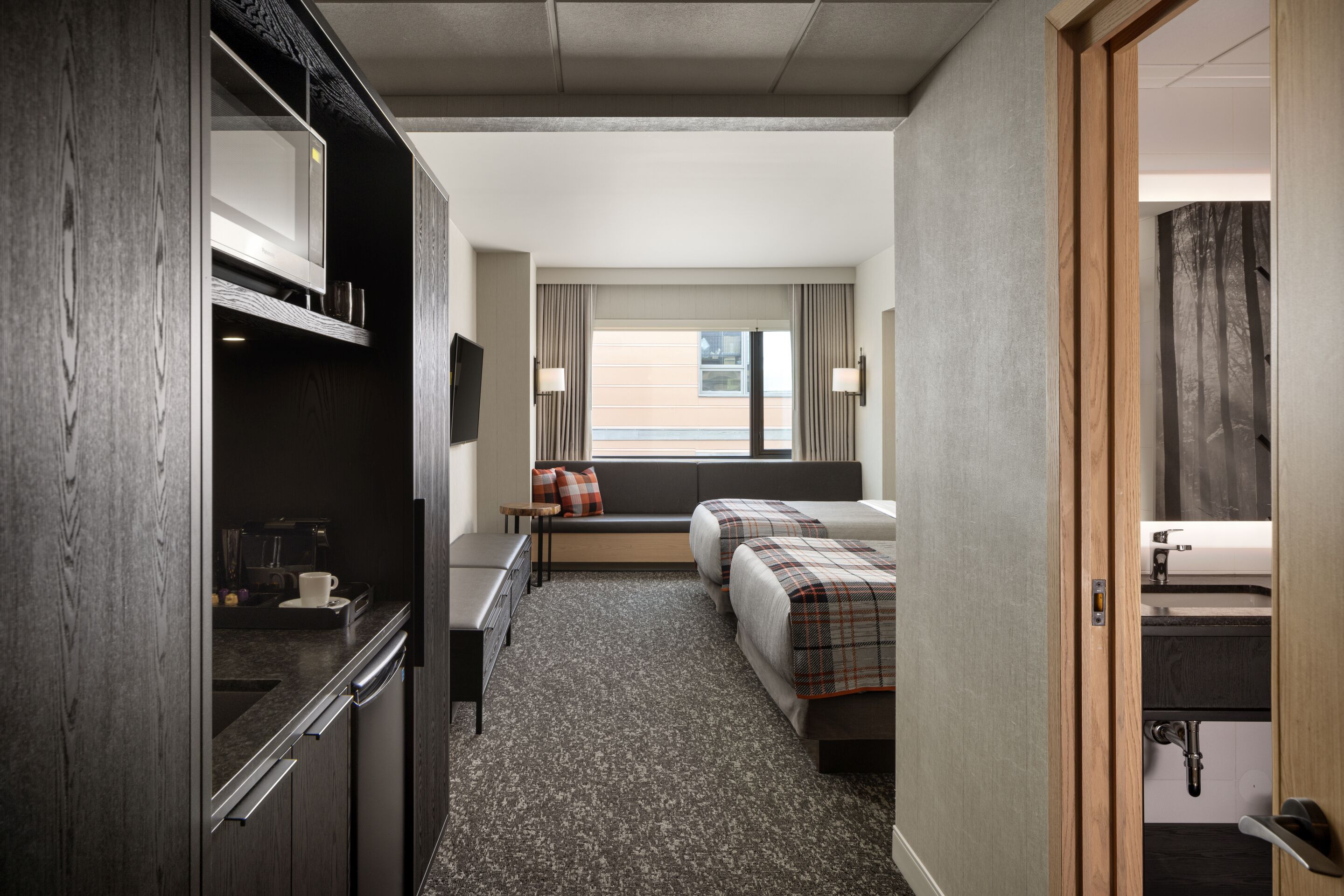 Habitación de hotel con kitchenette, cama con cubrecama escocés, ventana con cortinas y vista al exterior.