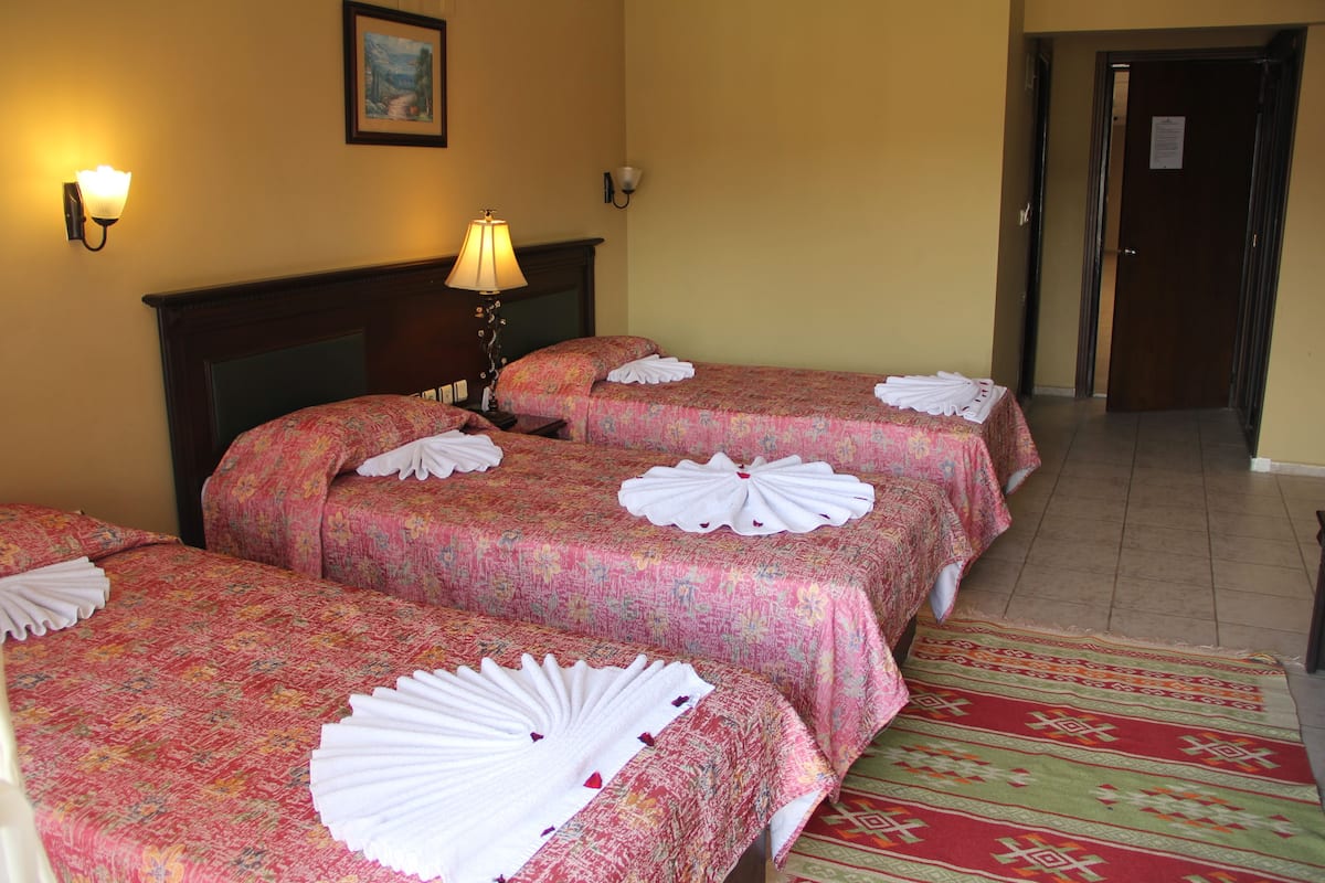 A hotel room with three beds, each with a floral bedspread and white towels. There is a lamp on the nightstand, a painting on the wall, and a door with a sign.