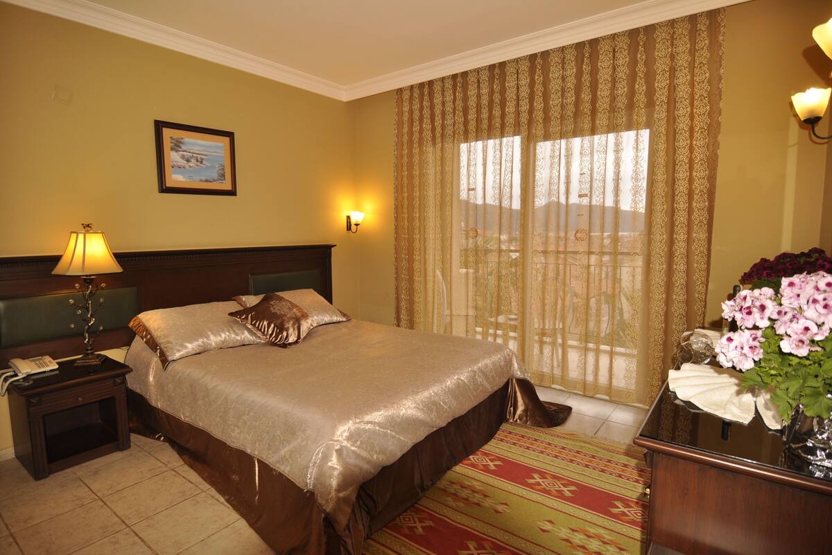 A hotel room with a large bed, a nightstand with a lamp, a framed picture on the wall, and a window with curtains.