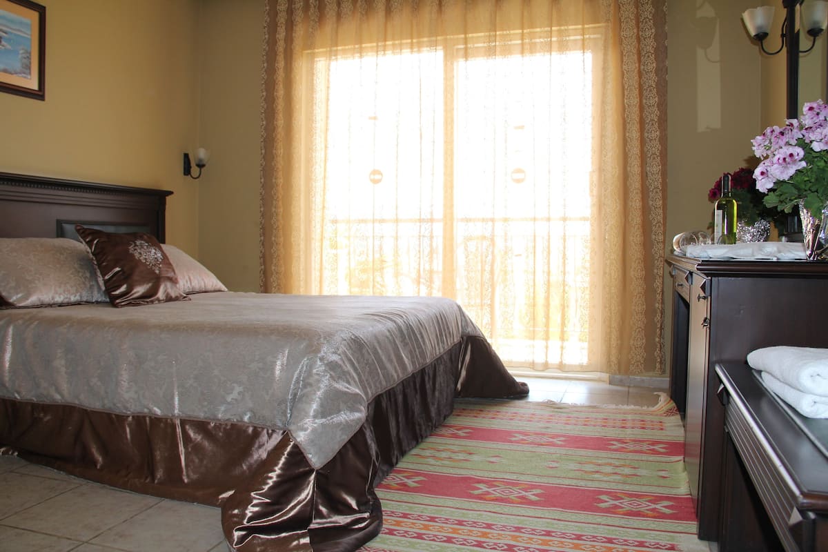 A neatly made bed with a brown bedspread, a headboard, and a nightstand with a vase of flowers and a bottle.