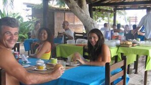 Un restaurant situé en bord de mer, doté d’une terrasse, abritée par une structure en bois, et dont les tables sont recouvertes de nappes vertes.