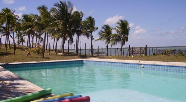 A swimming pool with clear blue water, surrounded by palm trees and a fence. - Pousada O Forte (Jandaira)