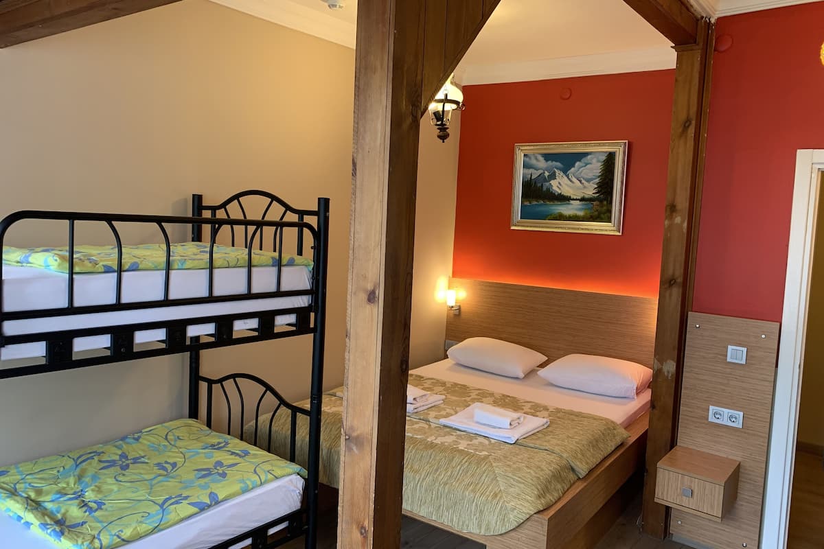 A room with two bunk beds and a single bed, wooden beams, a painting on the wall, and a nightstand with a lamp.