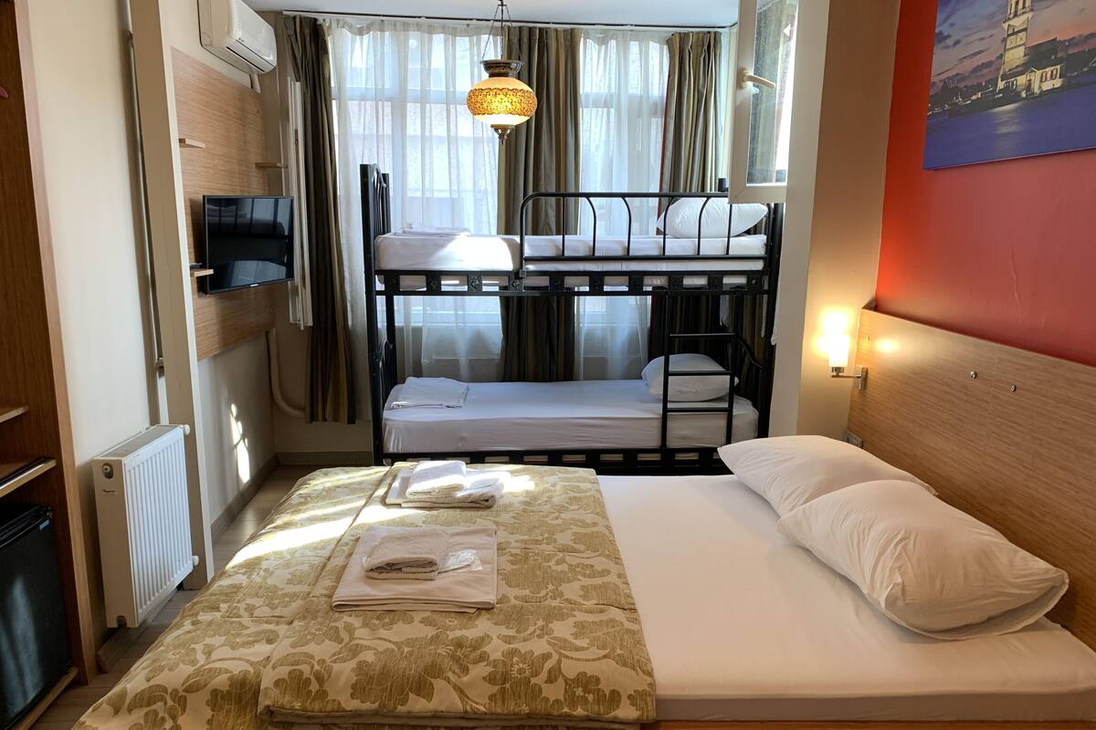 A hotel room with a bunk bed, a single bed with a floral bedspread, a radiator, a television, and a painting of a lighthouse.