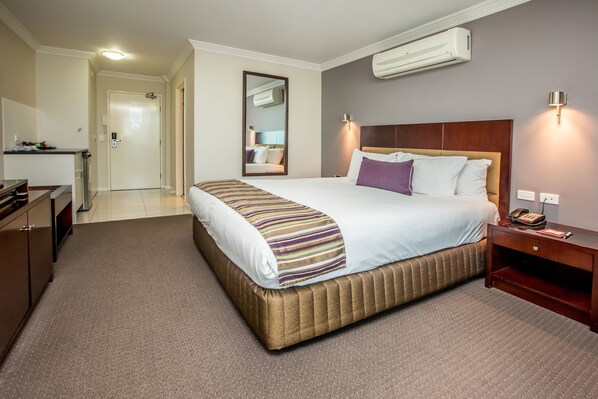 A hotel room with a large bed, a bedside table, a desk, and an air conditioning unit.