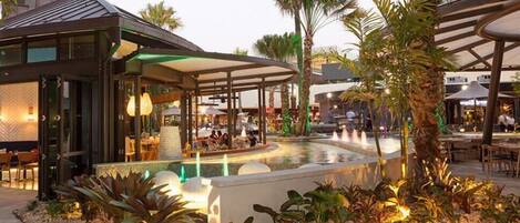A modern outdoor dining area with a pool, palm trees, and a covered seating area.