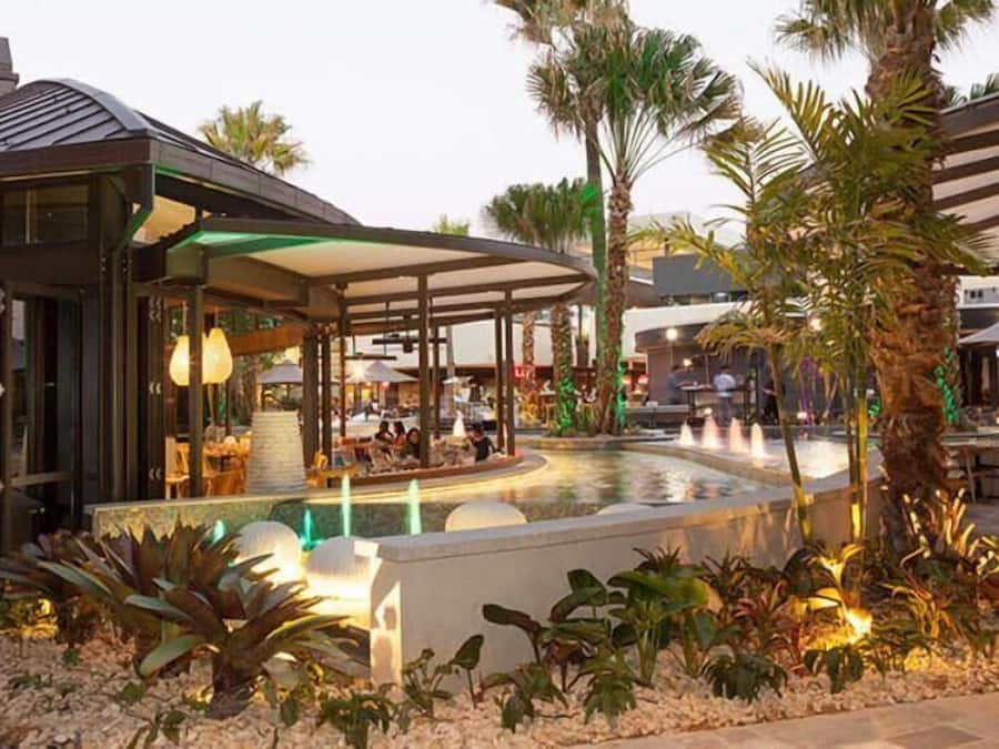Un área de comedor exterior moderna con piscina, palmeras y una zona de estar cubierta.