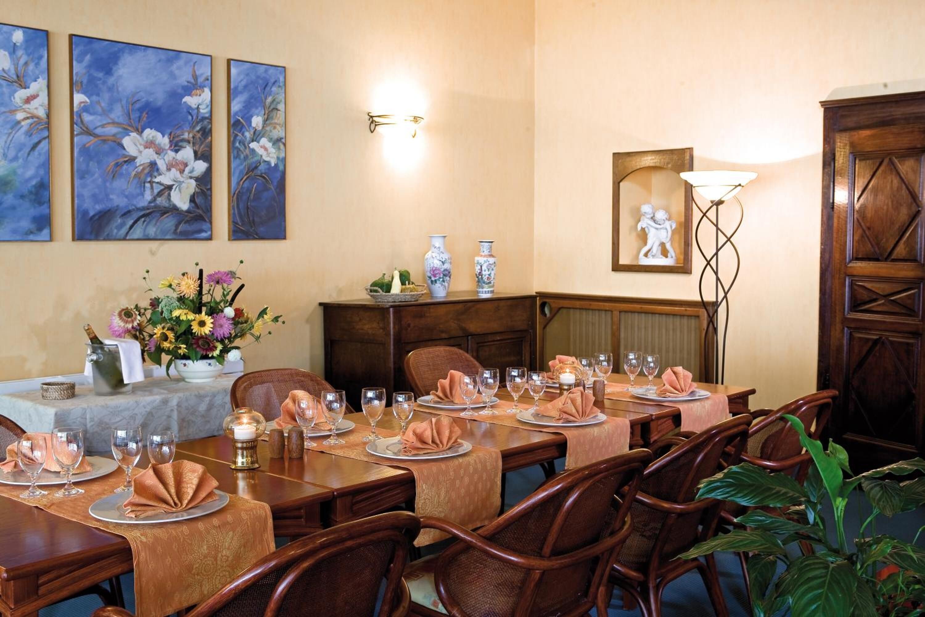 A dining area with a long table set for a meal, surrounded by chairs. There are floral decorations on the table, a vase with flowers, and a lamp.
