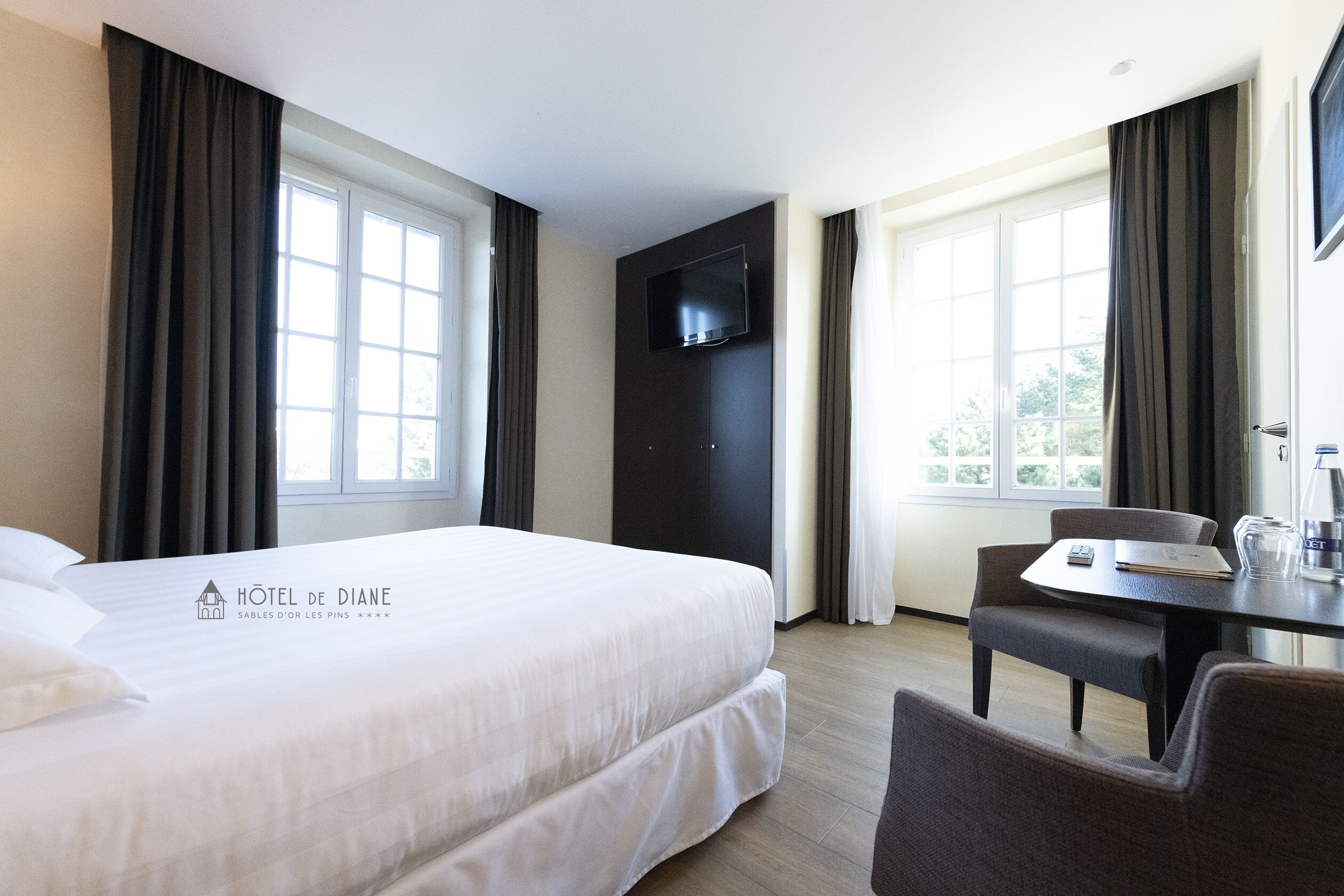 A hotel room with a large bed, a desk with a chair, a flat-screen TV mounted on the wall, and a window with curtains.