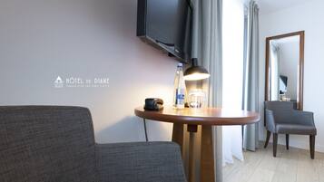 Una habitación de hotel moderna con una televisión de pantalla plana montada en la pared, una mesita redonda de madera con una lámpara, un vaso de agua y un teléfono. Un sillón gris y una silla están ubicados cerca de una ventana con cortinas.